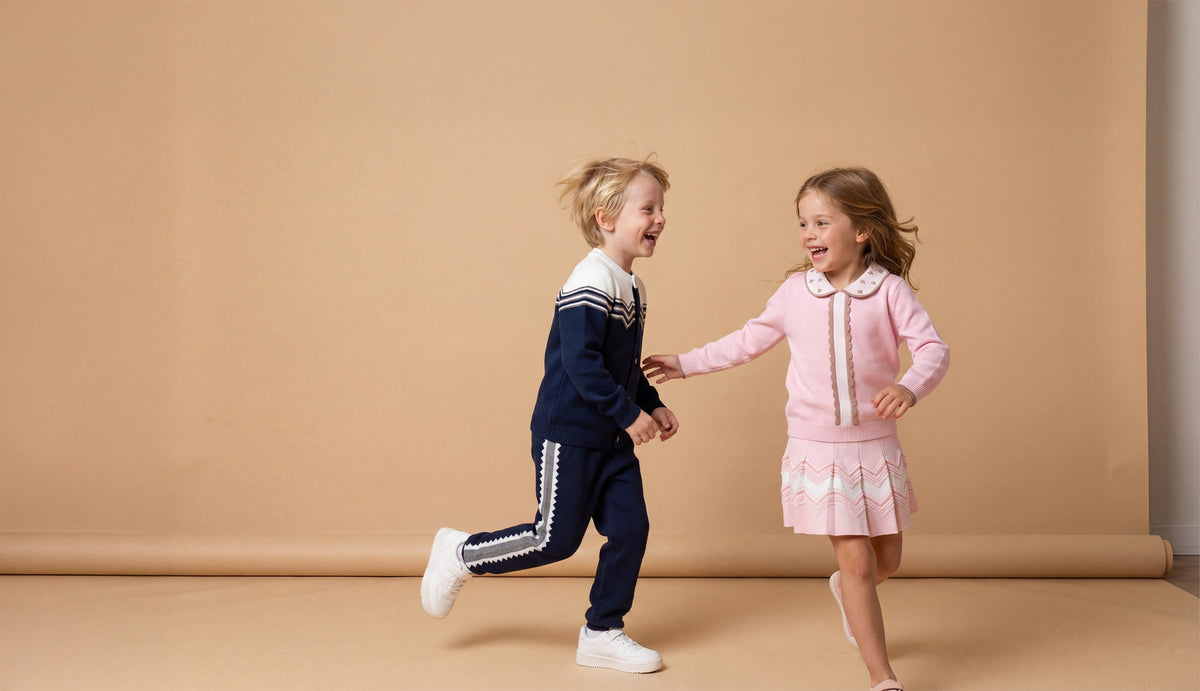 Two young children running and laughing while holding hands in a studio setting with a warm beige backdrop. The child on the left wears a navy blue knit outfit with white trim, and the child on the right wears a pink knit outfit with white trim.