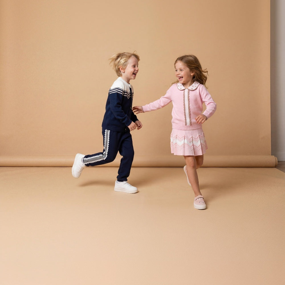 Two young children running and laughing while holding hands in a studio setting with a warm beige backdrop. The child on the left wears a navy blue knit outfit with white trim, and the child on the right wears a pink knit outfit with white trim.